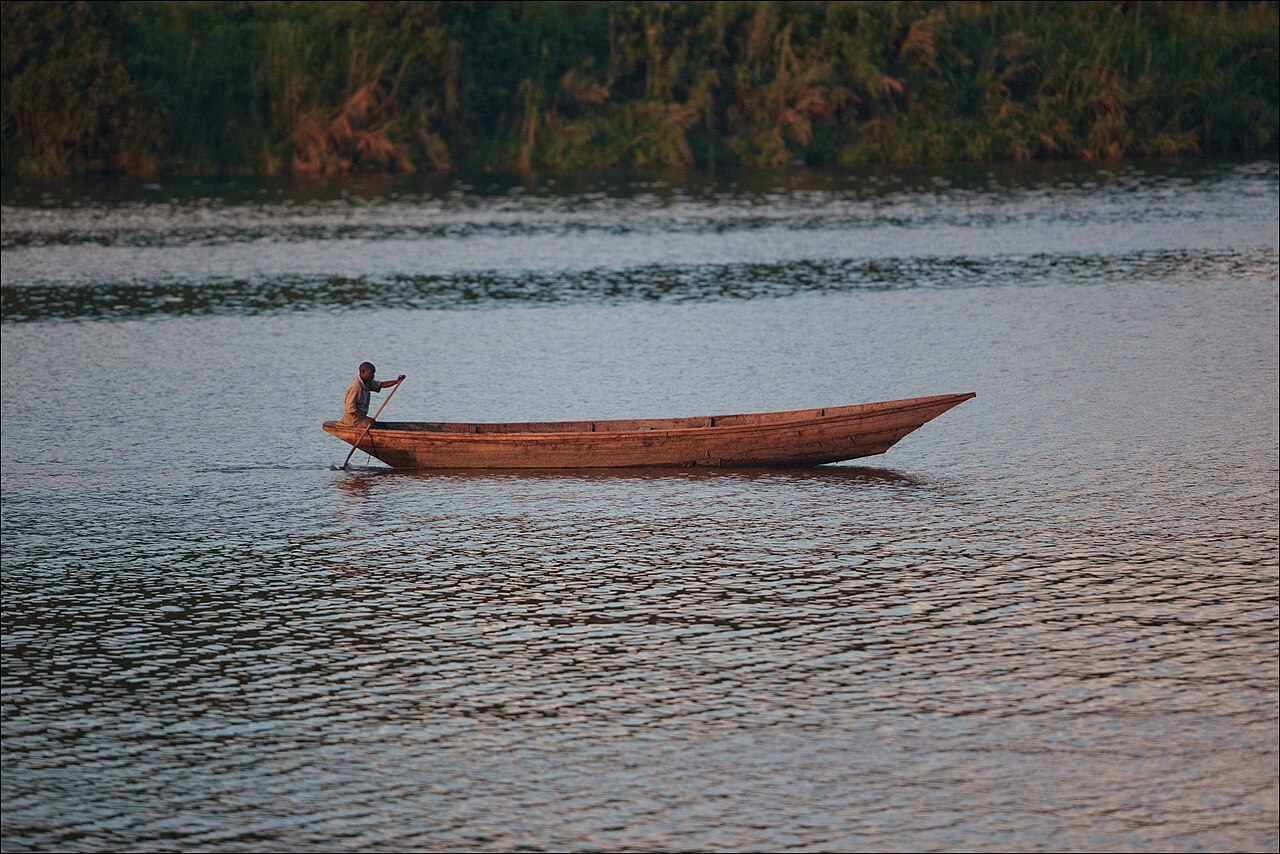 A man paddling a long wooden dugout canoe across the lake in early morning light